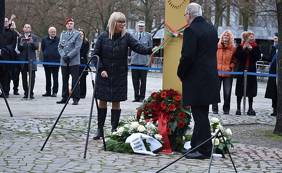 Zwei Menschen stehen vor einer Gedenkstele und öffnen eine Schleife.