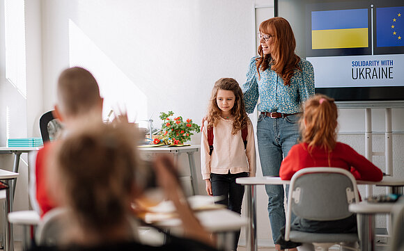 Ein Mädchen steht verschüchtert vor einer Schulklasse, die Lehrerin hat den Arm auf ihre Schulter gelegt