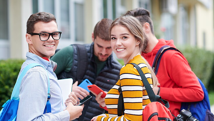 Vier Studenten unterhalten sich vor der Uni, zwei blicken über die Schulter in die Kamera
