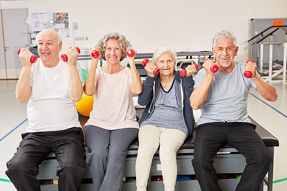 Vier Senioren sitzen in einem Fitnessraum auf einer Bank und stemmen mit ihren Armen kleine Hanteln. Sie lächeln dabei.