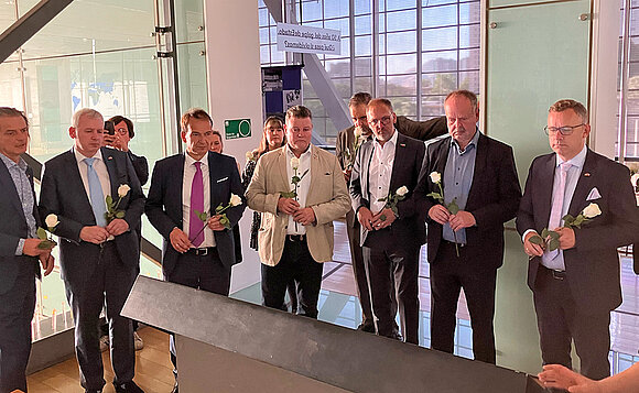 Männer mit Rosen in der Hand stehen vor einem Medientisch und folgen Erklärungen des Museumsguides.