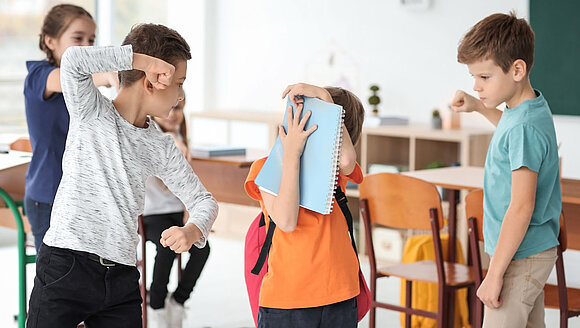 In einem Klassenzimmer erheben zwei Jungs ihre Fäuste gegen ein Mädchen, das sich hinter einem Heft versteckt und wegduckt.