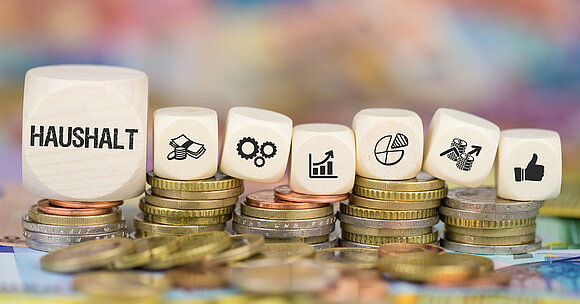 Geld und Würfel: Sie symbolisieren das Ringen um die Finanzierung der im Haushalt angestrebten Leistungen.