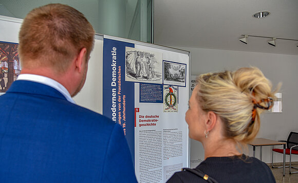 Über die Schultern zweier Besucher geht der Blick auf eine Ausstellungstafel.