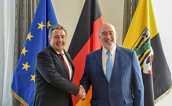 Landtagspräsident und Botschafter schütteln sich vor Fahnen die Hand.