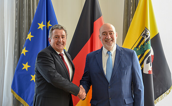 Landtagspräsident und Botschafter schütteln sich vor Fahnen die Hand.
