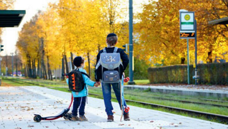 Zwei Schulkinder mit Kippa und Rucksäcken stehen wartend an einer Straßenbahnhaltestelle.
