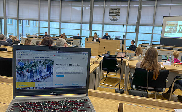 Im Vordergrund steht ein Laptop auf einem Tisch, im Hintergrund ist der Plenarsaal während der Ausschusssitzung zu sehen.