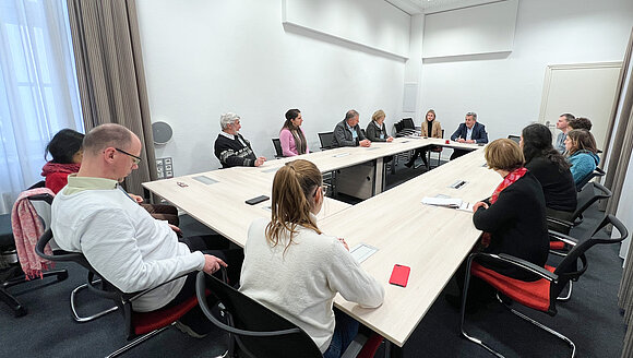 ltlsa/stb Menschen sitzen sich an einem rechteckigen Tisch gegenüber und unterhalten sich.
