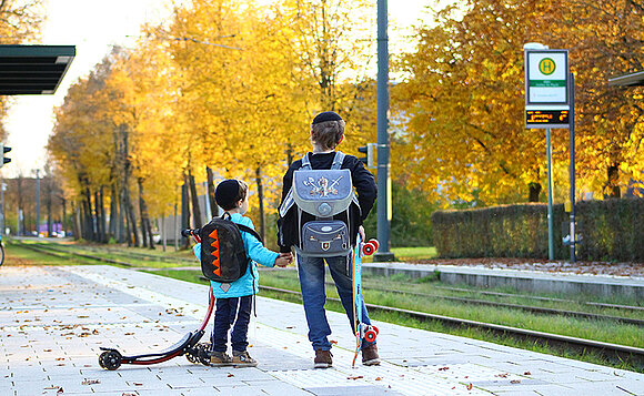 Zwei Schulkinder mit Kippa und Rucksäcken stehen wartend an einer Straßenbahnhaltestelle.