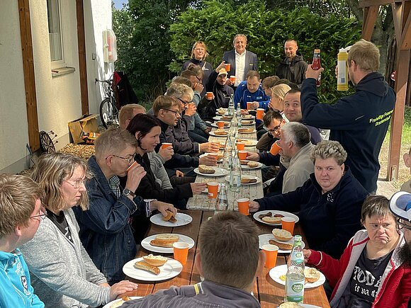 An einem langen gedeckten Tisch sitzen Jugendliche und Erwachsene, kurz vorm Essen.