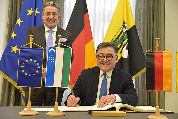 ltlsa/stb Botschafter sitzt am Tisch und schreibt etwas ins Gästebuch. Hinter ihm stehen Fahnen und der Landtagspräsident.