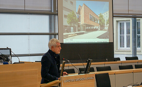 Architekt Ralf Niebergall steht am Rednerpult, hinter ihm ist eine Powerpoint-Präsentation zu sehen.