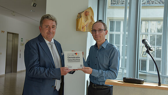 ltlsa/stb Zwei Männer halten gemeinsam eine Plakette in den Händen.