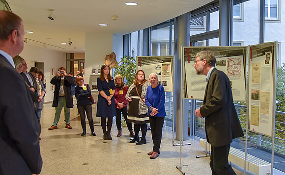 Gäste der Ausstellung hören zwischen den Ausstellungstafeln stehend einem Redner zu.