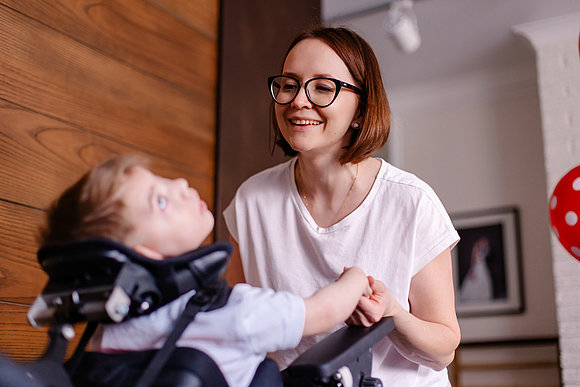 Kleinkind mit Mehrfachbehinderung im Rollstuhl wird von einer Frau fröhlich angelächelt.