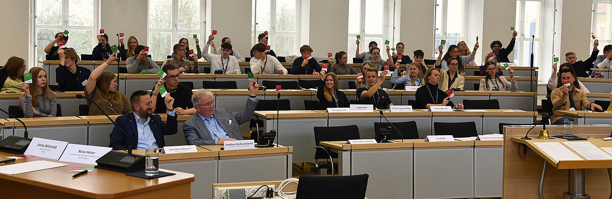 Jugendliche sitzen im Plenarsaal und heben Karten mit Pro und Kontra hoch