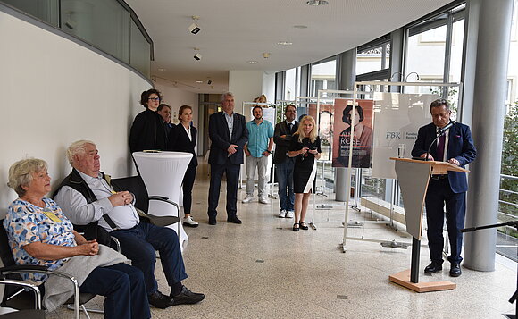 Gäste der Ausstellungseröffnung stehen und sitzen im Halbrund